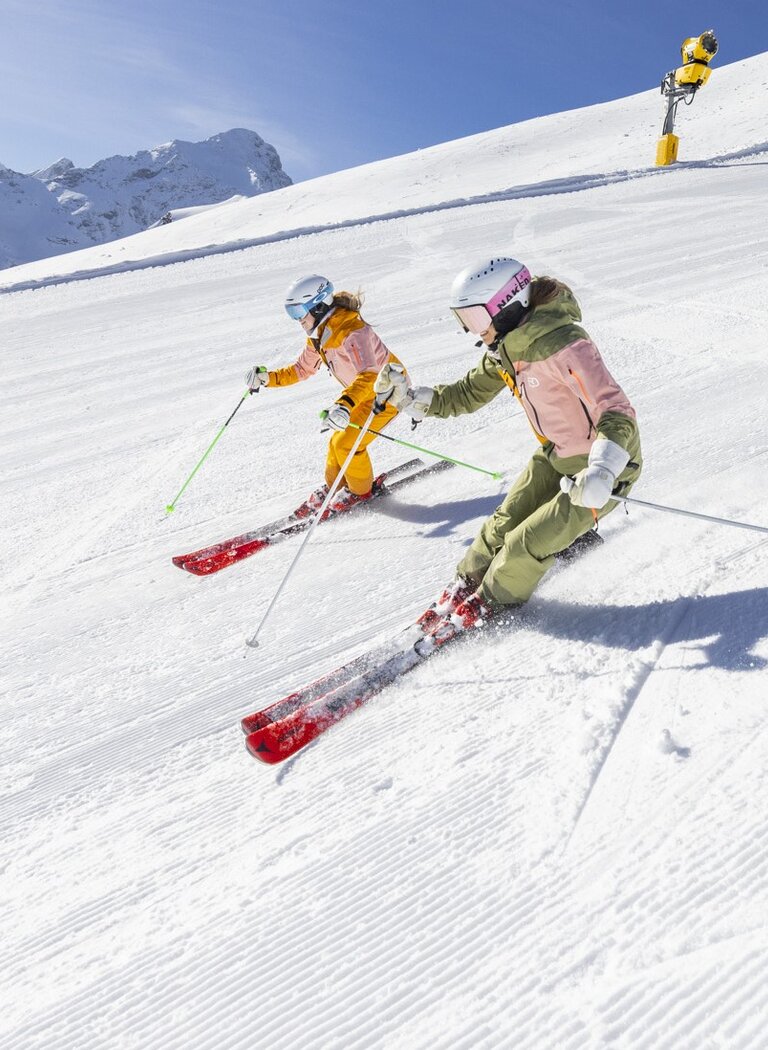 Zwei Frauen carven synchron auf frisch präparierter Skipiste bei Sonnenschein, umgeben von verschneiten Bergen und blauem Himmel | © Hauser Kaibling Seilbahn- und Liftges.m.b.H. & Co KG