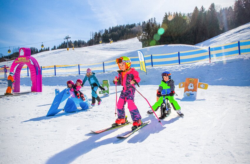 Kinder beim Skifahren und Spielen im Wollis Kinderparadies | © Hauser Kaibling Seilbahn- und Liftges.m.b.H. & Co KG Kinder fahren Ski und Laufrad im bunten Wollis Kinderparadies mit Parcours, Figuren und Zauberteppich bei Sonnenschein | © Hauser Kaibling Seilbahn- und Liftges.m.b.H. & Co KG