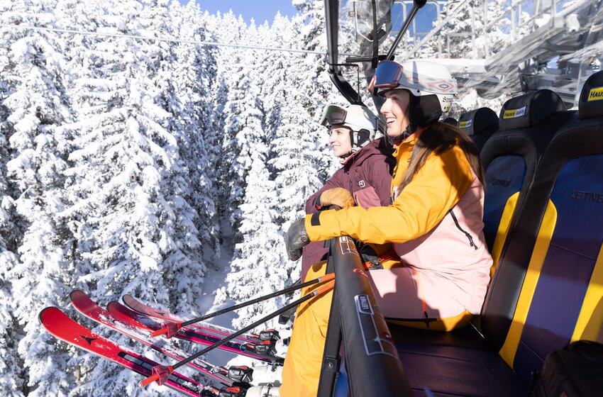 Two skiers wearing helmets sit on chairlift above densely snow-covered pine trees | © Hauser Kaibling Seilbahn- und Liftges.m.b.H. & Co KG