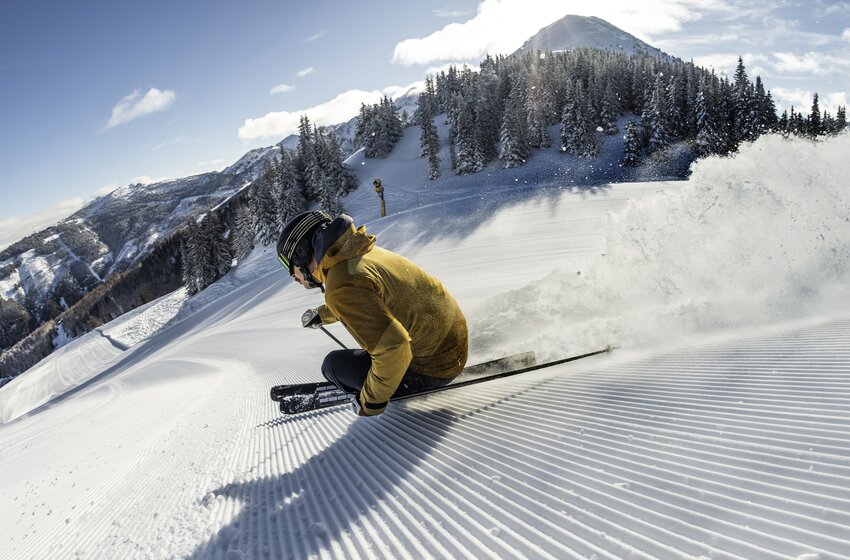 A skier makes a deep carving turn on a freshly groomed slope with snow-covered mountains and fir trees behind. | © Mirja Geh